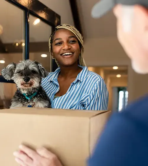 A smiling pet parent holding her small dog and accepting a delivery box at the door